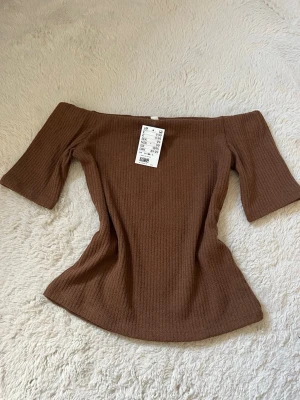 Brun ribbad offshoulder topp - Säljer en brun ribbad offshoulder topp från H&M. Toppen har trekvartsärm och är figurnära med en snygg, croppad passform. Perfekt att styla med höga jeans eller kjol för en trendig look. Jag brukar ha S eller XS och den passar mig jätte bra.