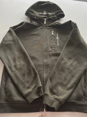 Olivgrön hoodie från Sail Racing - Olivgrön hoodie från Sail Racing med dragkedja framtill, bröstficka med zip och logga på bröstet. Insidan är fodrad med mjuk teddy för extra värme. Huva med dragsko och ribbade muddar vid ärmslut och nederkant. Perfekt för chill dagar eller när du vill ha något bekvämt.