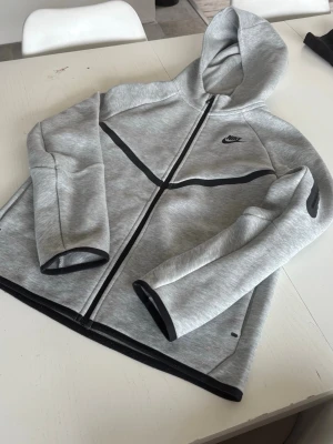 Grå Nike Tech Fleece hoodie - Snygg grå Nike Tech Fleece hoodie med dragkedja och svarta detaljer. Hoodien har en modern passform, huva och Nike-logga på bröstet. Tillverkad i mjukt och stretchigt material som känns riktigt skönt. Perfekt för dig som gillar sportig och clean stil.