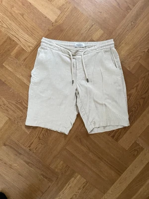 Beige linneshorts med snörning - Säljer ett par ljusbeige shorts från dressmann  i linne med elastisk midja och snörning. Shortsen har två sidofickor och en bakficka, perfekt för en avslappnad och stilren sommarlook.