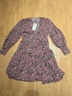 Blommig omlottklänning från Only - Kort omlottklänning från Only med blommigt mönster. Klänningen har långa ärmar, volangdetaljer och knyt i midjan💕