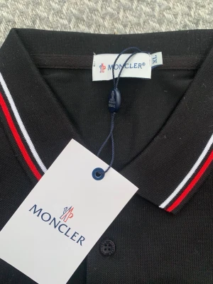 Svart Moncler pikétröja XL - Klassisk svart pikétröja från Moncler med vit och röd rand på kragen. Tröjan har knappslå med tre knappar och Moncler-logga broderad på bröstet. Perfekt för en stilren och sportig look.