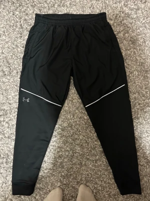 Svarta träningsbyxor Under Armour - Säljer ett par svarta träningsbyxor från Under Armour med vita reflexdetaljer över låren och logga på vänster ben. Byxorna har resår i midjan, smal passform och muddar vid bensluten. Perfekta för löpning eller gym. Materialet är lätt och andas. Storlek L men passar som M/L.