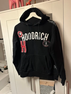 Svart hoodie från Hoodrich - Svart hoodie från Hoodrich med stora vita bokstäver på bröstet, röda och vita patchar samt broderad H-detalj. Klassisk känguruficka framtill och justerbar huva med snören. Tjockt och mjukt material, perfekt för en streetwear-look. Storlek M.