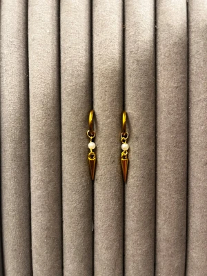 Guldörhängen med pärlor och koner - Snygga örhängen i guldfärgad metall med små vita pärlor och hängande konformade detaljer. Örhängena har en modern och edgy look med minimalistisk design. Perfekta för dig som vill ha något unikt och trendigt i smyckeskrinet.