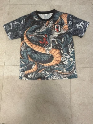 T-shirt med drakmönster och Japan-emblem - Unik t-shirt med stort, detaljerat drakmönster i blått, beige och grått. Svart krage och Japan-emblem på bröstet. Perfekt för dig som gillar streetwear och asiatiska influenser.