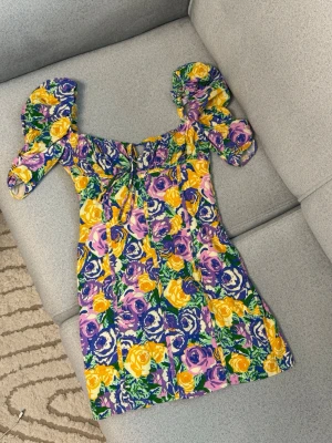 Blommig kortklänning från Zara - Färgstark kortklänning från Zara med puffärmar och blommigt mönster i gult, lila, grönt och vitt. Klänningen har en söt snörning framtill och dragkedja i sidan. Perfekt för dig som gillar statement-plagg och vill sticka ut i sommar!