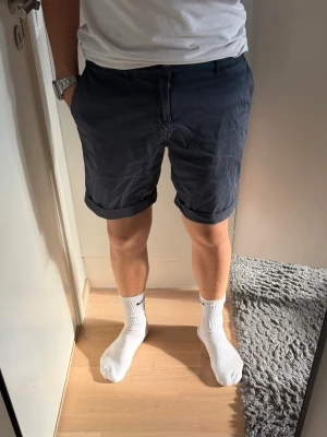 Mörkblå shorts med uppvik  - Snygga mörkblå shorts i bomull med uppvikta benslut och klassisk passform. Shortsen har sidofickor och bälteshällor, perfekta för en avslappnad stil. Passar till t-shirt eller skjorta för en enkel look.