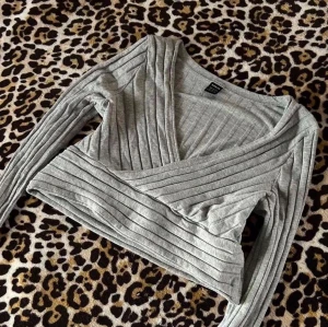 Grå ribbad omlottopp - Snygg grå croppad omlottopp med ribbad struktur och långa ärmar. Djup v-ringning och tight passform som framhäver midjan. Perfekt att styla med höga jeans eller kjol.