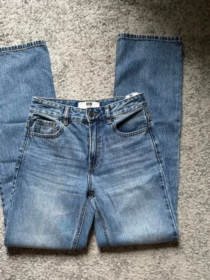 Blå raka jeans från STR - Säljer ett par klassiska blå jeans från STR med rak passform och fem fickor. Jeansen har en snygg tvättad look och är tillverkade i denim med en patch bak i midjan. Perfekta för dig som gillar en enkel och tidlös stil.
