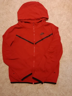 Röd Nike hoodie med dragkedja - Snygg röd hoodie från Nike med hel dragkedja framtill och svart Nike-logga på bröstet. Hoodien har svarta detaljer längs ärmslut och nederkant samt en huva. Tillverkad i ett mjukt och bekvämt material, perfekt för en sportig och avslappnad stil.