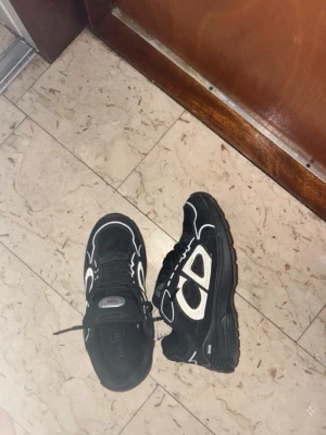 Svarta sneakers  - Säljer ett par svarta sneakers med vita detaljer och tjock sula. Skorna har en låg profil och är tillverkade i en mix av mocka och textil. Snygg kontrast mellan svart och vitt samt unika mönster på sidorna. Perfekta för dig som gillar streetwear och vill sticka ut.