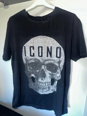Svart t-shirt med dödskalletryck ICONO - Cool svart t-shirt från ICONO med stort dödskalletryck framtill och glittriga detaljer. T-shirten har rund hals och korta ärmar. Perfekt för dig som gillar edgy och unik stil.
