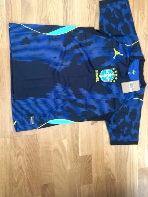 Blå Brasilien matchtröja  - Snygg blå och svart Brasilien matchtröja från Puma med gula detaljer och Brasil-logga på bröstet. Tröjan har korta ärmar, rund hals och coolt mönster över hela tyget. Perfekt för dig som älskar fotboll och vill sticka ut på planen.