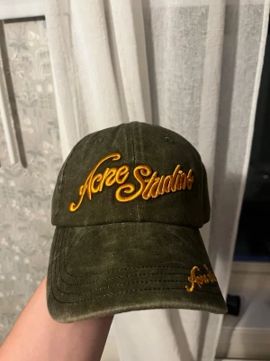 Olivgrön keps från Acne Studios - Snygg olivgrön keps från Acne Studios med broderad gul logga framtill och på skärmen. Klassisk böjd skärm och justerbar baksida. Tillverkad i bomullstyg med vintagekänsla. Perfekt accessoar för en cool streetstyle-look.