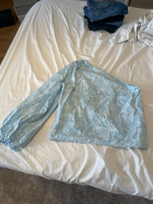 Blå off shoulder topp med bladmönster - Säljer en ljusblå off shoulder topp med ett vitt bladmönster. Toppen har en lång puffärm med resår vid ärmslutet och en rak passform. Perfekt för dig som vill ha en unik och trendig look. Materialet känns lätt och luftigt.
