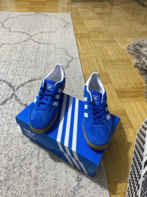 Blå Adidas Gazelle  - Snygga blå Adidas Originals sneakers i mocka med klassiska vita ränder på sidorna och vit insida. Skorna har en låg siluett och gummisula som ger en retrovibe. Perfekta för dig som vill ha en stilren och sportig look.