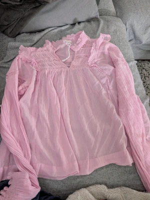 Ljusrosa blus med volangdetaljer stl 158-164 - Supersöt ljusrosa blus från Gina Tricot med volangkant vid kragen och spetsdetaljer upptill. Blusen har v-ringning, tunna långärmade ärmar och är gjord i ett tunt, skirt tyg som ger en romantisk vibe. Perfekt att styla med jeans eller kjol.