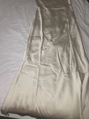 Champagnefärgad långklänning i satin - Elegant långklänning i champagnefärgad satin med slät och glansig yta. Klänningen har en enkel, rak siluett och är axelbandslös för en stilren look. Perfekt för dig som vill ha en minimalistisk och chic stil.