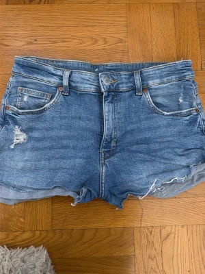 Blå denimshorts med slitningar - Snygga blå denimshorts med rå kant och slitna detaljer framtill. Klassisk femficksmodell med knapp och dragkedja. Passar perfekt till sommaren och har en avslappnad, loose fit. Inga synliga loggor men typisk jeanslook. Skulle säga att de passar storlek M också