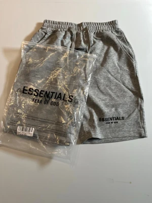 Grå shorts från Essentials Fear of God - Säljer ett par grå shorts från Essentials Fear of God med snörning i midjan och fickor på sidorna. Klassisk logga tryckt på vänster ben. Sköna och stilrena shorts i mjuk bomullsblandning, perfekta för sommaren.
