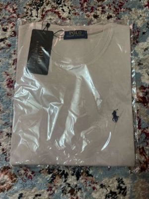 Beige t-shirt från Polo Ralph Lauren - Snygg beige t-shirt från Polo Ralph Lauren med klassisk rund hals och den ikoniska blå loggan broderad på bröstet. Tillverkad i mjuk bomull för en skön känsla. Perfekt basplagg till garderoben.