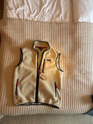Beige fleeceväst från Patagonia - Snygg beige fleeceväst från Patagonia med marinblå detaljer och röda dragkedjor. Västen har tre fickor med dragkedja och klassisk ståkrage. Perfekt för lager-på-lager och outdoor-stil. Materialet är mjuk fleece som håller dig varm.