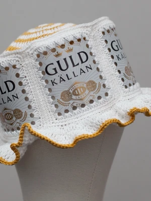 Virkad ölhatt Guldkällan - Unik virkad ölhatt med Guldkällan.  Virkad i 100% bomull. Passar upp till ca 60cm.  Vissa burkar kan ha mindre repor eller bucklor, vilket tyvärr kan uppstå under tillverkningen av hattarna. Insidan av burkarna slipas efter hålen tagits för att undvika vassa kanter. Därför kan det ibland finnas lite slipmärken, men det är inget som stör eftersom det är på insidan av hattarna.  Färger kan variera något beroende på skärminställningar.