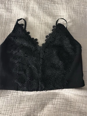 Svart spetsbody med smala axelband - Snygg svart fest topp med tunna axelband och v-ringning. Framsidan är dekorerad med detaljerad spets i blommönster som ger en elegant look. Perfekt att styla under en skjorta eller kavaj. Materialet känns mjukt och följsamt mot huden.