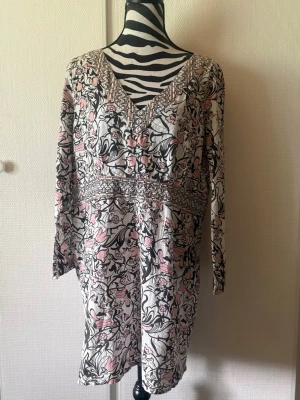 Blommig långärmad kortklänning - Säljer en söt kortklänning med blommigt mönster i vitt, svart och ljusrosa. Klänningen har v-ringning, långa ärmar och ett dekorativt band i midjan. Perfekt för dig som gillar romantiska och feminina plagg.