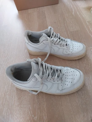 Vita Nike Air Force 1 sneakers - Klassiska vita Nike Air Force 1 obs storlek 43 sneakers i lågt utförande. Skorna har en stilren design med perforerad tåbox, vita skosnören och sula i gummi. Tillverkade i läder med textilfoder för extra komfort. Perfekta för dig som gillar tidlös streetstyle. Tyget vid hälarna har gått sönder se bild. Lagas lätt med en lapp. Finns ett litet hål i sulan längst bak på ena skon se bild annars i mycket bra skick. 