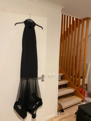 Svart långklänning med meshdetalj - Elegant svart långklänning med hög hals och ärmlös design. Klänningen har en unik meshdetalj nedtill som ger en modern och stilren look. Perfekt för dig som vill sticka ut på festen med en sofistikerad och trendig outfit.