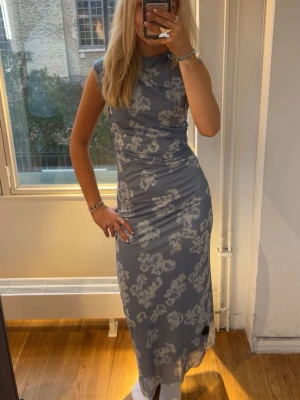 Blå blommig långklänning  - Snygg blå långklänning med vitt blommönster. Klänningen har en figurnära passform och är ärmlös, vilket ger en modern och stilren look. Perfekt för sommarens alla tillfällen. Enbart använd en gång! Nypris ca 1200kr