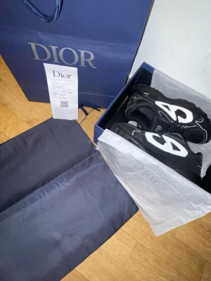 Svarta Dior B30 sneakers - Säljer ett par svarta Dior B30 sneakers med stora vita CD-loggor på sidorna. Skorna har en chunky sula, meshpaneler och detaljer i läder. Snygg och modern design som sticker ut, perfekta för dig som gillar exklusiva märken och streetwear.