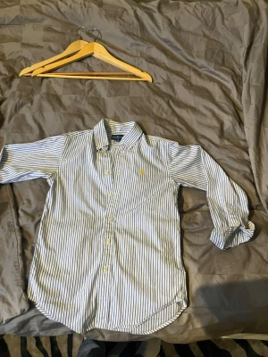 Randig skjorta från Ralph Lauren - Säljer en klassisk vit skjorta med blå ränder från Ralph Lauren. Skjortan har lång ärm, knappar framtill och den ikoniska loggan broderad på bröstet. Tillverkad i 100% bomull för en skön och luftig känsla.
