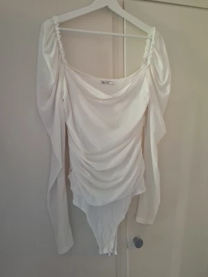 Vit omlott bodysuit från NLY One - Säljer en vit bodysuit från NLY One med långa ärmar och draperad omlott-design. Toppen har en bred, rund ringning och är tillverkad i ett lätt, stretchigt material som ger en snygg passform. Perfekt för att styla med jeans eller kjol. Obs finns nån fläck (se bild) den är inte genomskinlig dä materialet är tjockare vid bröstdelen