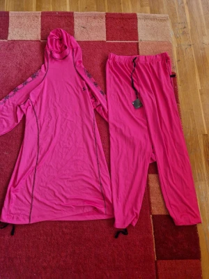 Rosa set med huva och byxor - Tvådelat set i starkt rosa färg, består av en långärmad tunika med huva och svarta sömdetaljer samt matchande byxor med resår och dragsko i midjan. Tunikan har diskret mönster på ärmarna. Materialet ser ut att vara mjukt och stretchigt, perfekt för avslappnade dagar. Från Dubai