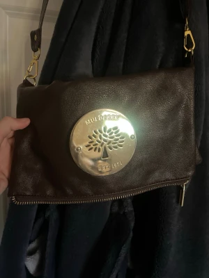 Mörkbrun axelväska från Mulberry - Snygg mörkbrun axelväska i äkta läder från Mulberry med stort guldfärgat metallmärke framtill och präglad logga. Väskan har justerbar axelrem och dragkedja längst ner. Perfekt för dig som gillar klassisk och stilren design.