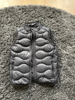Svart dunväst Peak Performance - Snygg svart dunväst från Peak Performance med vågigt quiltat mönster och hög krage. Västen har dragkedja framtill och två sidofickor. Perfekt för lager på lager och håller dig varm under kyliga dagar.