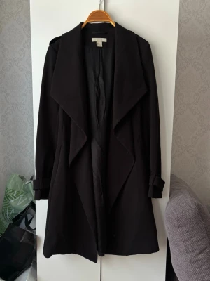 Svart kappa - Svart kappa / trenchcoat från H&M i strl 32 med knytskärp i midjan och dekorativa axelklaffar. Jackan har långa ärmar med spännen vid handlederna och en fin ryggdetalj!❤️