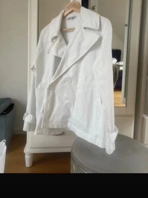 Vit dubbelknäppt rock - Stilren vit rock med dubbelknäppning och bred krage. Rocken har stora vita knappar, två sidofickor och justerbara remmar vid ärmsluten. Klassisk och enkel design som passar till många olika looks. Det är XS/S