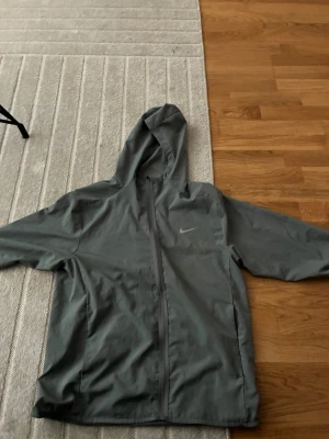 Grå vindjacka från Nike med huva - Snygg grå vindjacka från Nike med hel dragkedja och huva. Jackan har två sidofickor och diskret Nike-logga på bröstet. Perfekt för blåsiga dagar och enkel att matcha med olika outfits.