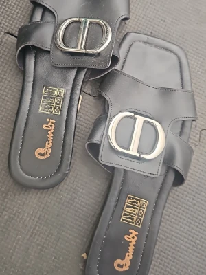 Svarta sandaler med metallspänne Bambi - Stilrena svarta sandaler från Bambi med fyrkantig tå och bred rem över foten. Dekorerade med ett stort, silverfärgat metallspänne framtill. Tillverkade i konstläder med guldfärgad logga på innersulan. Perfekta för en trendig och avslappnad look.