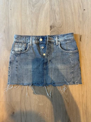 Blå jeanskjol från Zara, XS - Säljer en blå jeanskjol från Zara i storlek XS. Kjolen har rå kant nertill, klassiska fem fickor och knappar framtill. Perfekt för dig som gillar en avslappnad och trendig stil.