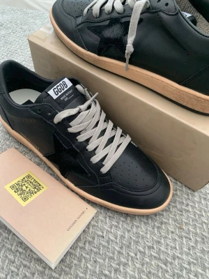Svarta Golden Goose Ball Star sneakers - Svarta Golden Goose Ball Star sneakers i läder med svart stjärndetalj på sidan, grå skosnören och beige sula. Klassisk låg modell med perforerad tå och GGDB-logga på plösen. Snygg och stilren design som passar till många outfits.