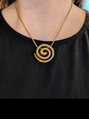 Guldigt halsband med spiralhänge - Unikt halsband med ett stort spiralformat hänge i guldtonad metall. Kedjan är tunn och matchar hängets färg, vilket ger ett coolt och iögonfallande uttryck. Perfekt för dig som gillar statement-smycken och vill sticka ut med din stil. Halsbandet är av kirurgiskt stål och tål värme samt vatten.