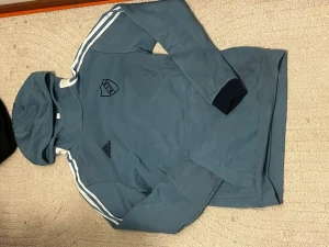 Blå DIF hoodie från Adidas - Snygg blå hoodie från Adidas med DIF-logga på bröstet och klassiska vita ränder på axlarna. Tröjan har huva och en stor magficka. Perfekt för dig som vill representera Djurgården med stil. Materialet känns mjukt och bekvämt.