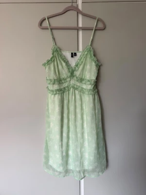 Ljusgrön sommarklänning  - Ljusgrön klänning med vitt blommönster och fina volangdetaljer. Klänningen har justerbara axelband, v-ringning och en luftig passform som gör den perfekt för sommarens alla tillfällen. Klänningen har även en vit underkjol.  Endast använd vid ett tillfälle sedan dess hängt i garderoben.