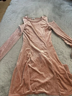 Rosa sammetsklänning med lång ärm - Supersnygg rosa sammetsklänning med lång ärm och axelpartier som är öppna. Klänningen har en mjuk och glansig finish som ger en lyxig känsla. Perfekt för dig som vill sticka ut med en unik och trendig look.