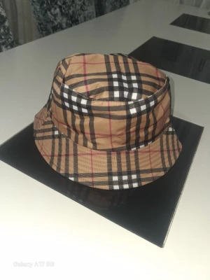 Burberry hat - Snygg bucket hat med klassiskt rutigt mönster i beige, svart, vitt och röda detaljer. Hatten har en mjuk form, perfekt för att lyfta din outfit med en trendig accessoar.